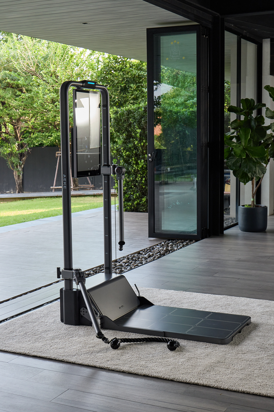 Compact home strength-training machine with a touchscreen and cable attachments in a bright room beside glass doors.